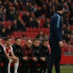 Een trainer langs de zijlijn tijdens een wedstrijd van Ajax, met focus op de bank en de sfeer in het stadion.