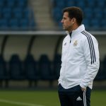 Zijaanzicht van een trainer op het trainingsveld van Real Madrid, met spelers op de achtergrond zonder herkenbare gezichten.