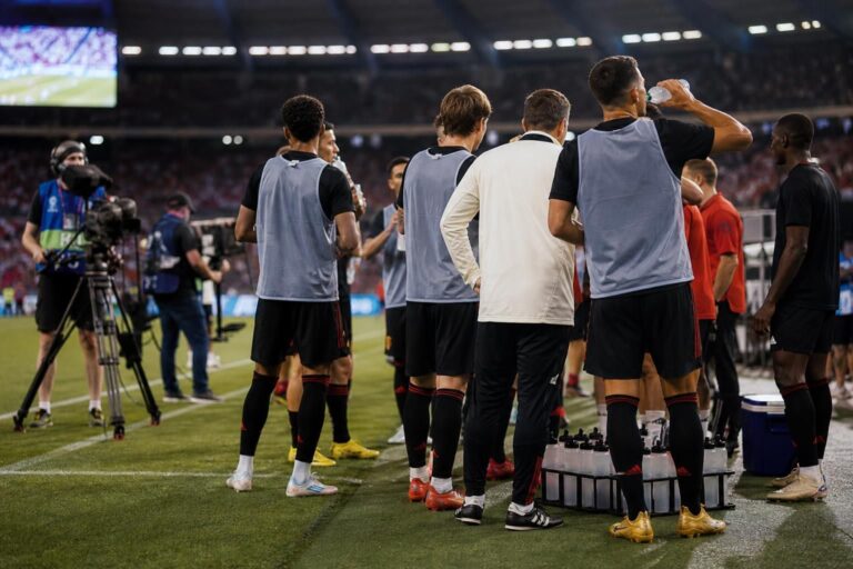 Een hydration break langs de zijlijn tijdens een voetbalwedstrijd met tv-camera’s op de achtergrond