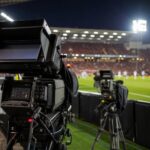 Tv-camera's langs de zijlijn in een Belgisch voetbalstadion tijdens een avondwedstrijd