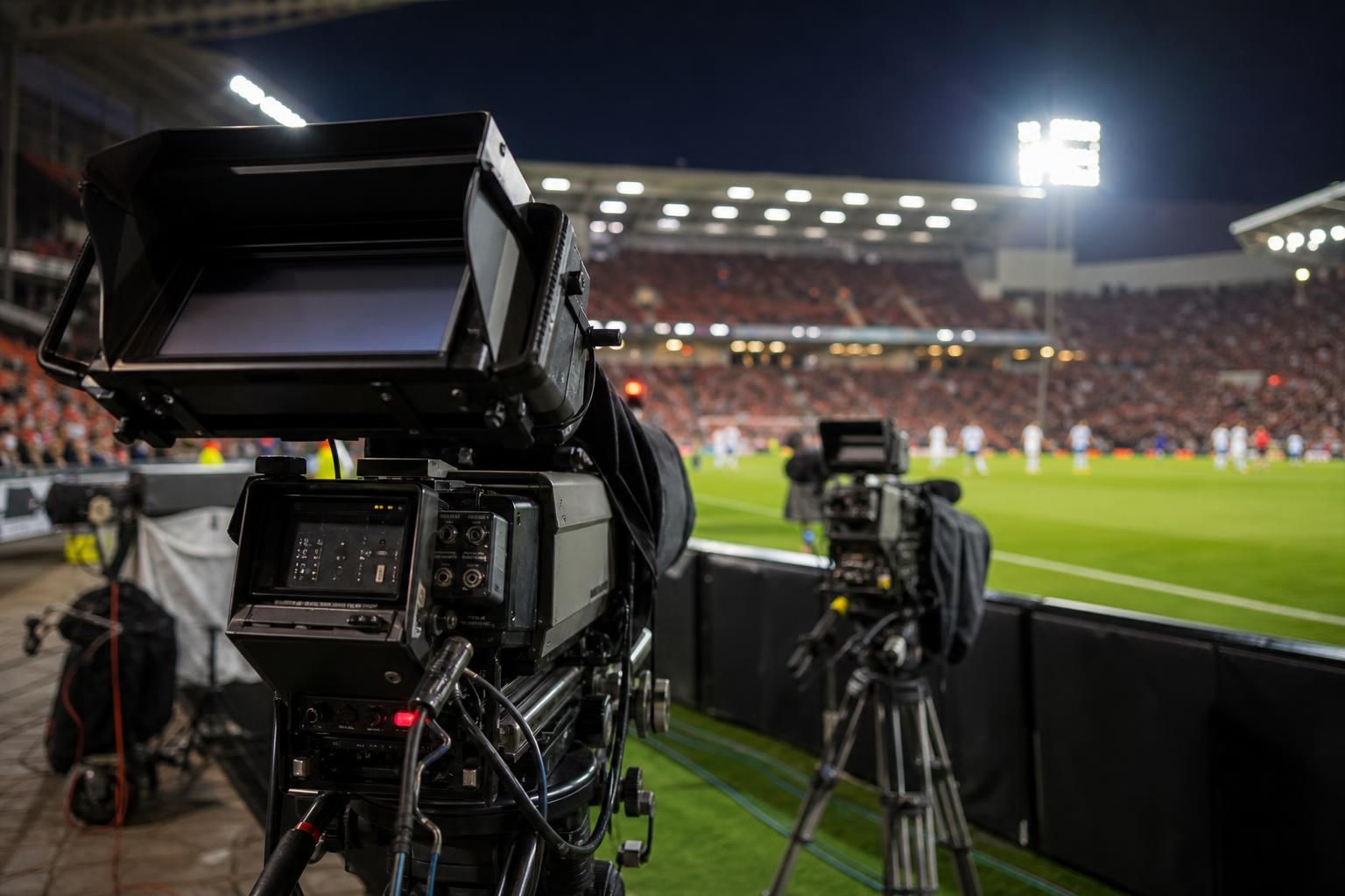 Tv-camera's langs de zijlijn in een Belgisch voetbalstadion tijdens een avondwedstrijd