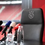 Persmoment rond een trainersdossier met microfoons en stadionachtergrond zonder herkenbare gezichten
