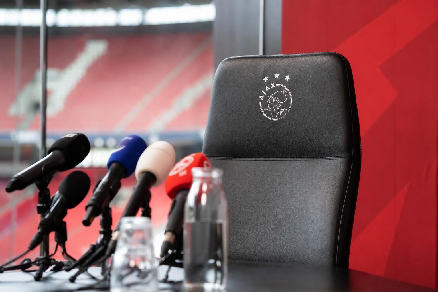 Persmoment rond een trainersdossier met microfoons en stadionachtergrond zonder herkenbare gezichten