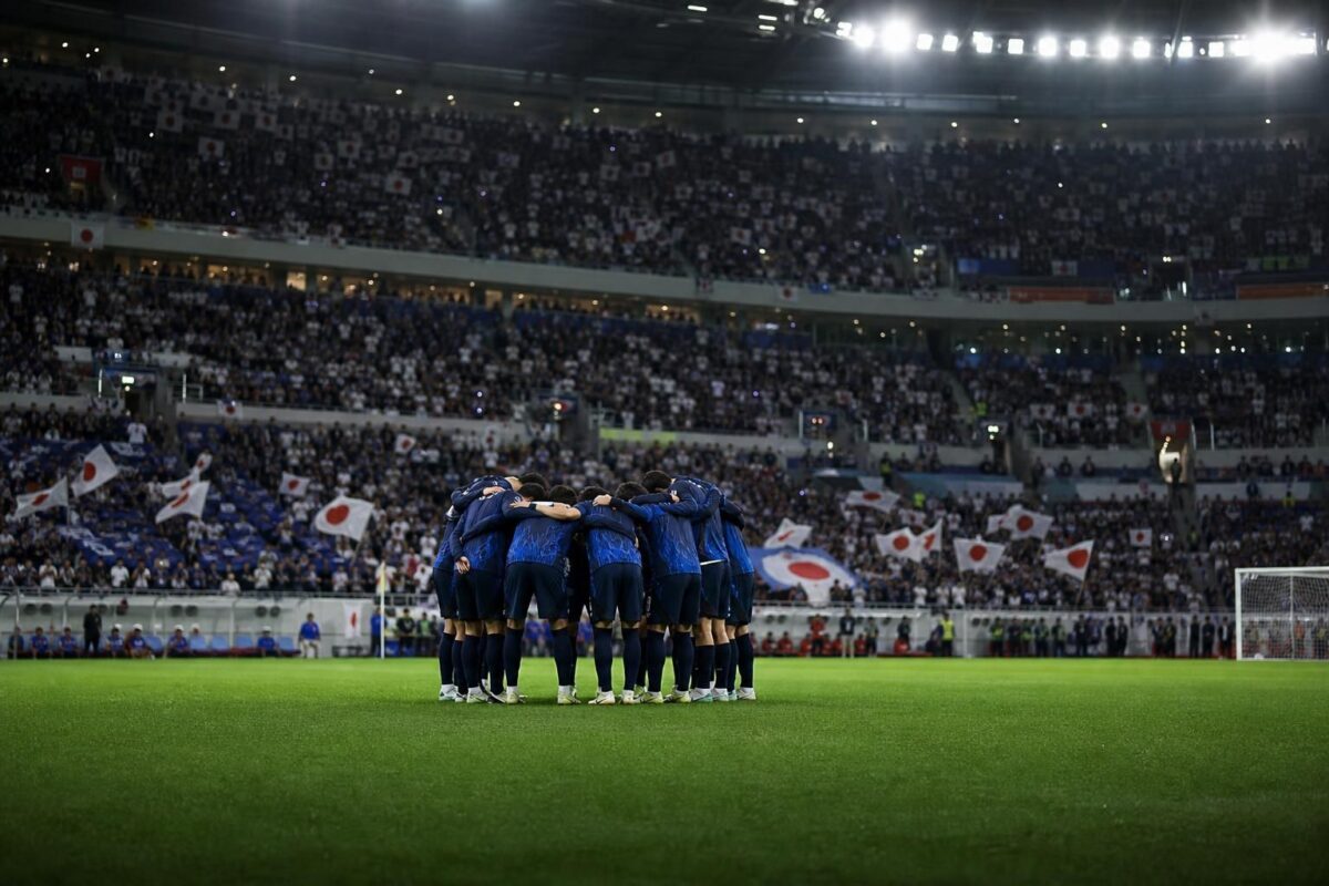 Japanse spelers vormen een kring voor een interland met supporters en stadionlicht op de achtergrond.