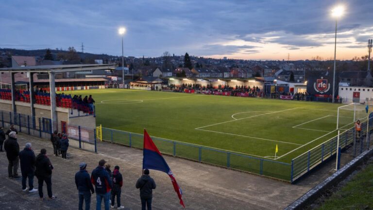 Het veld en de compacte tribunes van Stade de Rocourt, met rood-marineblauwe accenten die verwijzen naar RFC Liège.