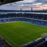 De blauw-witte tribunes en het volledige speelveld van KRC Genk in de Cegeka Arena, met supporters op de achtergrond.