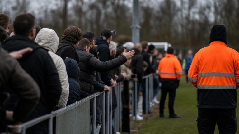Supporters en spelers rond een Vlaams voetbalveld in een geladen sfeer zonder herkenbare gezichten
