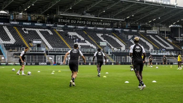 Breed wedstrijddagbeeld van het stadion van Sporting Charleroi met een opwarmende spelersgroep.