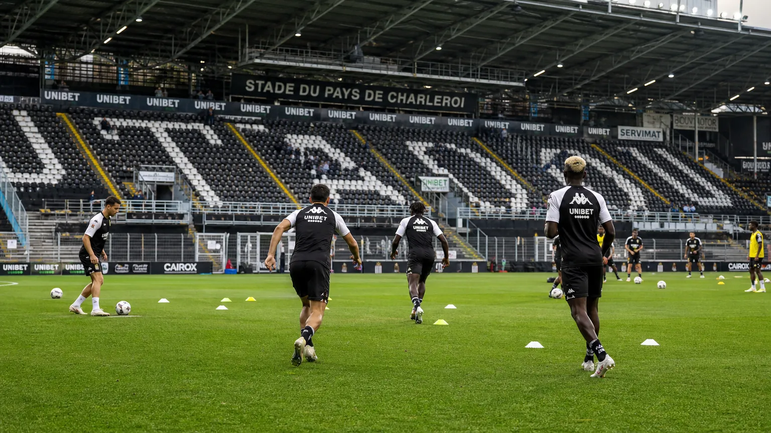 Breed wedstrijddagbeeld van het stadion van Sporting Charleroi met een opwarmende spelersgroep.