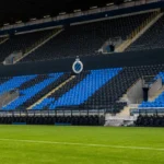 Een lege blauw-zwarte tribune met een Club Brugge-embleem aan de rand van het veld.