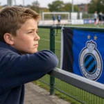 Een jonge supporter kijkt langs een jeugdvoetbalveld naar een Club Brugge-vlag.