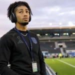 Nathan-Dylan Saliba in zwarte sportkleding aan de zijlijn van een voetbalstadion.