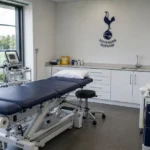Een lege medische onderzoeksruimte in een voetbalclub met een discreet Tottenham-embleem aan de wand.