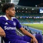 Nathan-Dylan Saliba zit in een stadiontribune in een paars Anderlecht-tenue.