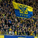 STVV-supporters juichen en zwaaien met een geel-blauwe vlag op een volle tribune.