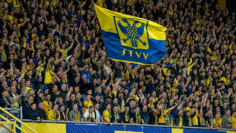 STVV-supporters juichen en zwaaien met een geel-blauwe vlag op een volle tribune.
