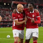Twee spelers van Royal Antwerp FC vieren samen tijdens een wedstrijd, met een speler van KRC Genk op de achtergrond.