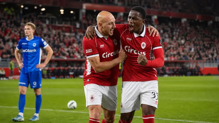 Twee spelers van Royal Antwerp FC vieren samen tijdens een wedstrijd, met een speler van KRC Genk op de achtergrond.