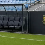 Een lege technische bank langs een voetbalveld met een discreet Zuid-Afrikaans embleem op de stadionwand.