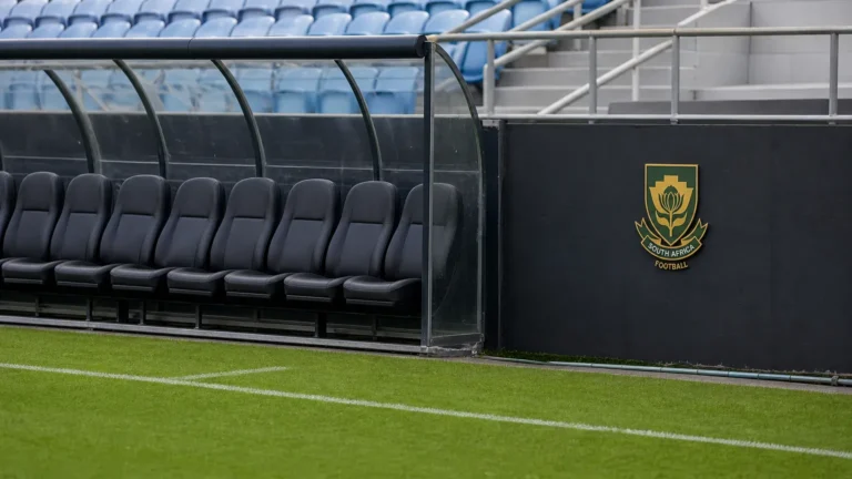 Een lege technische bank langs een voetbalveld met een discreet Zuid-Afrikaans embleem op de stadionwand.