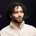 César Huerta in een lichte jas in een indoor voetbalomgeving.