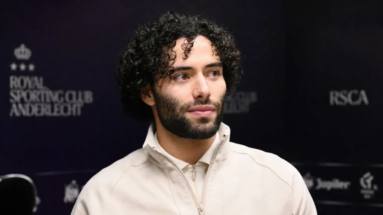 César Huerta in een lichte jas in een indoor voetbalomgeving.