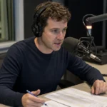 Een radiopresentator spreekt in een voetbalstudio naast een microfoon en notities.