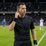 Een scheidsrechter communiceert tijdens een avondwedstrijd in een Belgisch voetbalstadion.