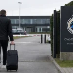 Een niet-herkenbare man met reistas wandelt langs een Club Brugge-bord aan het trainingscomplex.