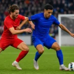 Twee voetballers in rode en blauwe onbedrukte tenues vechten om de bal nabij het strafschopgebied.