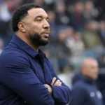 Troy Deeney kijkt aandachtig toe langs de zijlijn van een voetbalstadion.