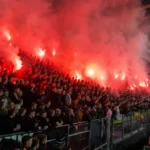 Supporters in een voetbalstadion terwijl rode fakkels veel rook verspreiden.
