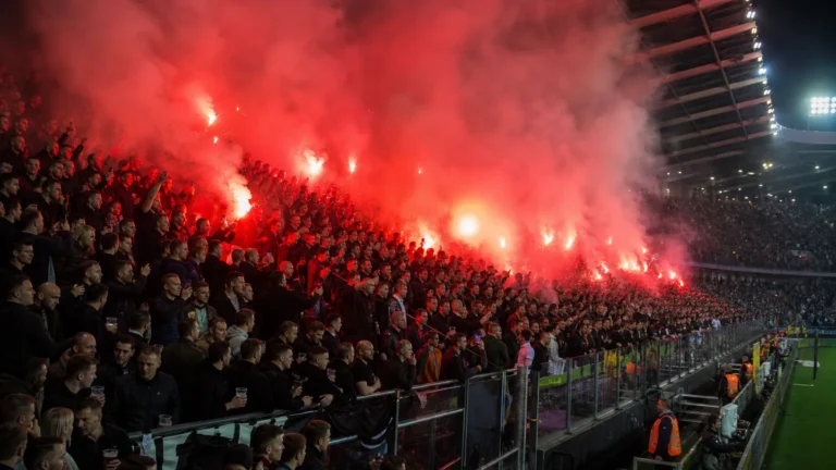 Supporters in een voetbalstadion terwijl rode fakkels veel rook verspreiden.