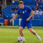 Liam Delap in blauw Chelsea-trainingskleding met de bal op het veld.