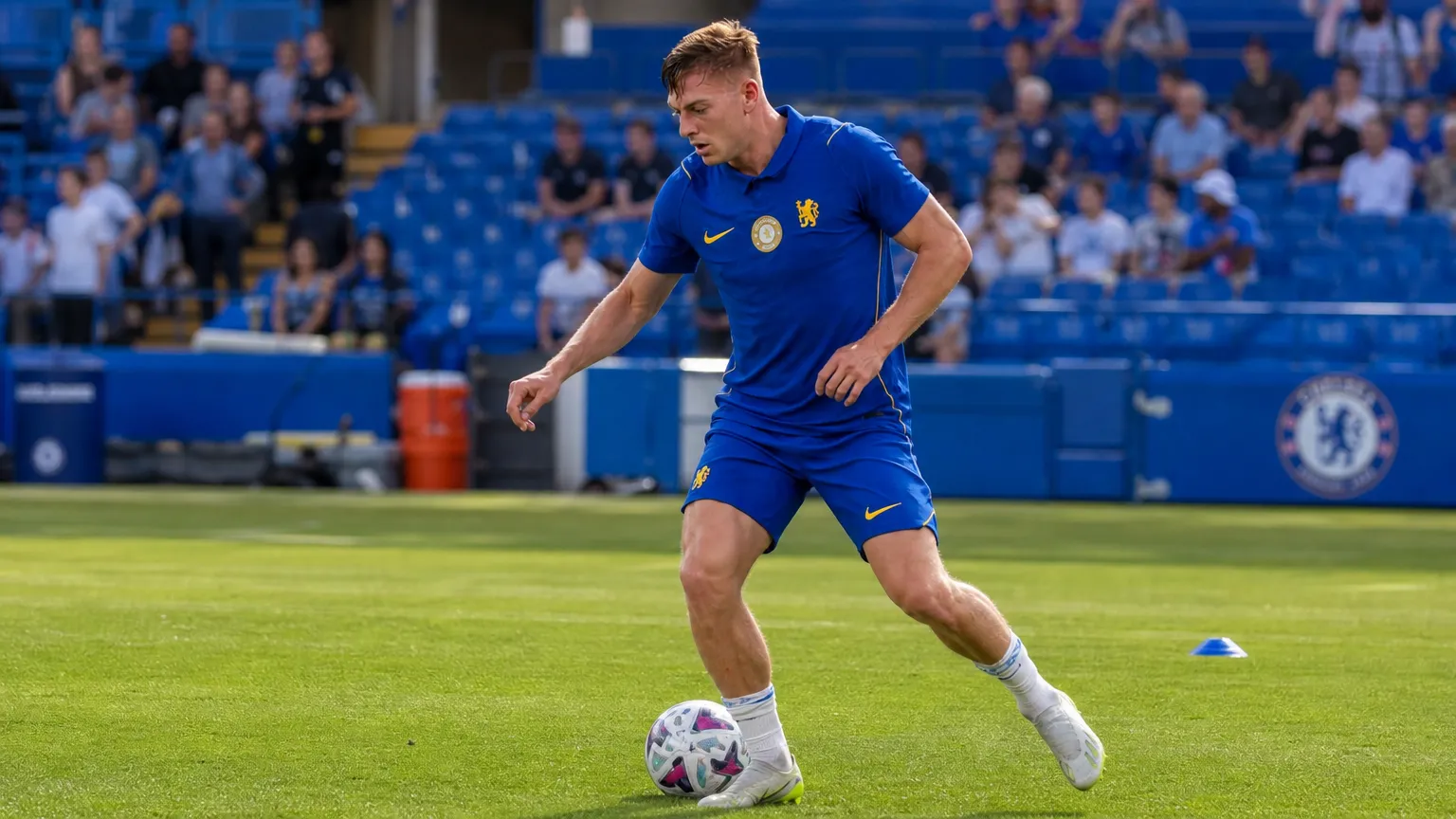 Liam Delap in blauw Chelsea-trainingskleding met de bal op het veld.