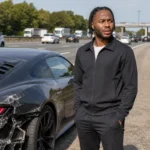 Raheem Sterling staat naast een beschadigde sportwagen langs een snelweg.