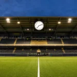 Een verlicht voetbalveld met een discreet Bodø/Glimt-bord en een stadionklok voor een bekeravond.