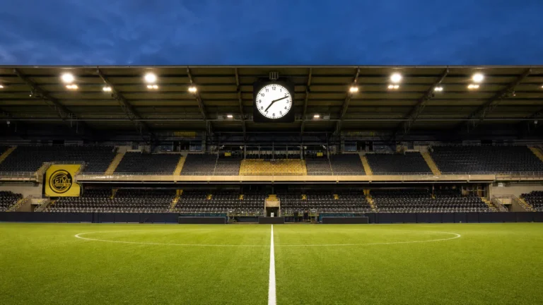 Een verlicht voetbalveld met een discreet Bodø/Glimt-bord en een stadionklok voor een bekeravond.