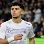 Yassin Belkhdim in een wit voetbalshirt aan de rand van het veld voor een wedstrijd.