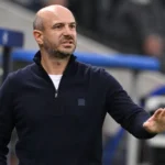 Een coach geeft geconcentreerd instructies langs de zijlijn van een voetbalveld.