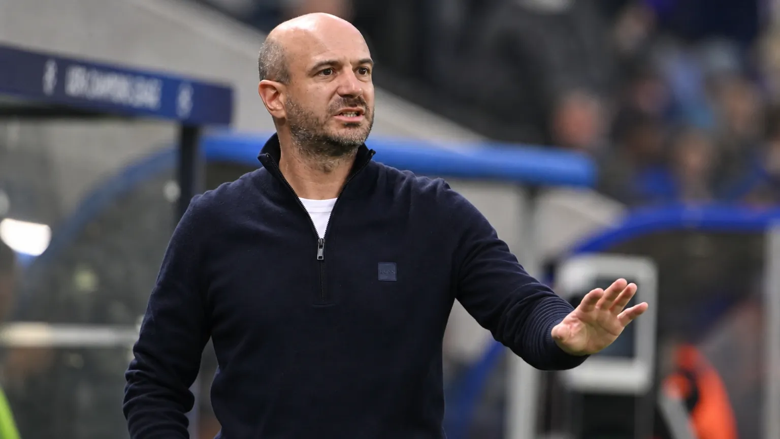 Een coach geeft geconcentreerd instructies langs de zijlijn van een voetbalveld.