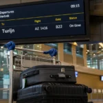 Vertrekbord met een vlucht naar Turijn naast een bagagetrolley in een luchthavenhal.