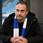 Sam Baro in zwarte jas en blauw-witte sjaal tijdens een interview.