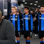 Frank Boeckx spreekt voor een microfoon met blauw-zwarte voetballers op de achtergrond.