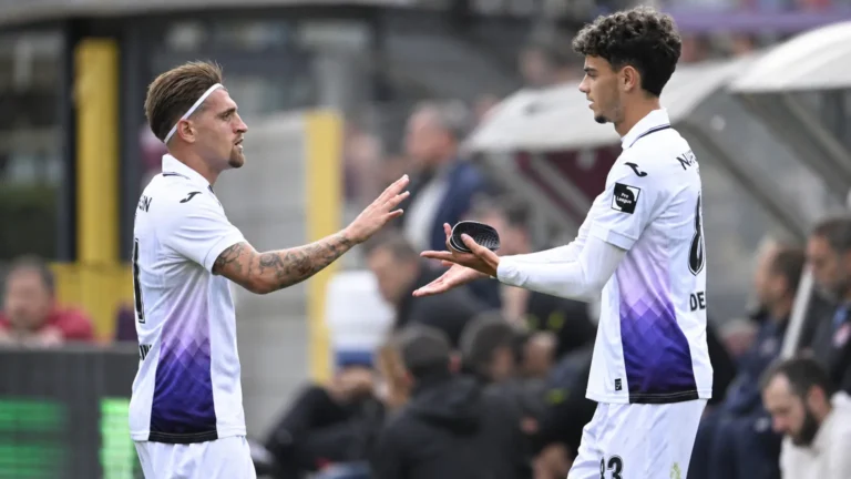 Twee voetballers in witte shirts met paarse accenten wisselen een aanvoerdersband uit.