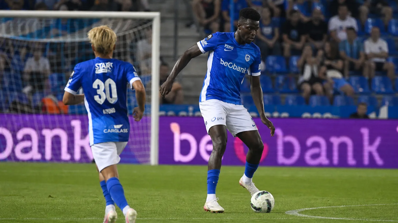 Twee voetballers in blauw-witte tenues tijdens een aanval in een Belgisch voetbalstadion.