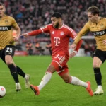 Een speler van Bayern München dribbelt met de bal terwijl twee spelers van Stuttgart achtervolgen.