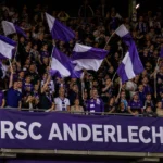 Anderlecht-supporters met paars-witte vlaggen in een volle tribune.