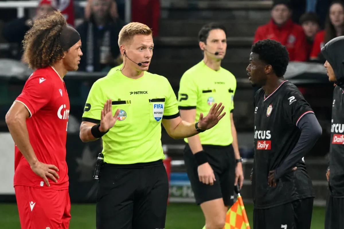 Een scheidsrechter spreekt met spelers in rode en donkere tenues tijdens een voetbalwedstrijd.