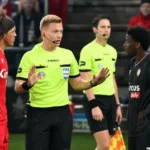 Een scheidsrechter spreekt met spelers in rode en donkere tenues tijdens een voetbalwedstrijd.